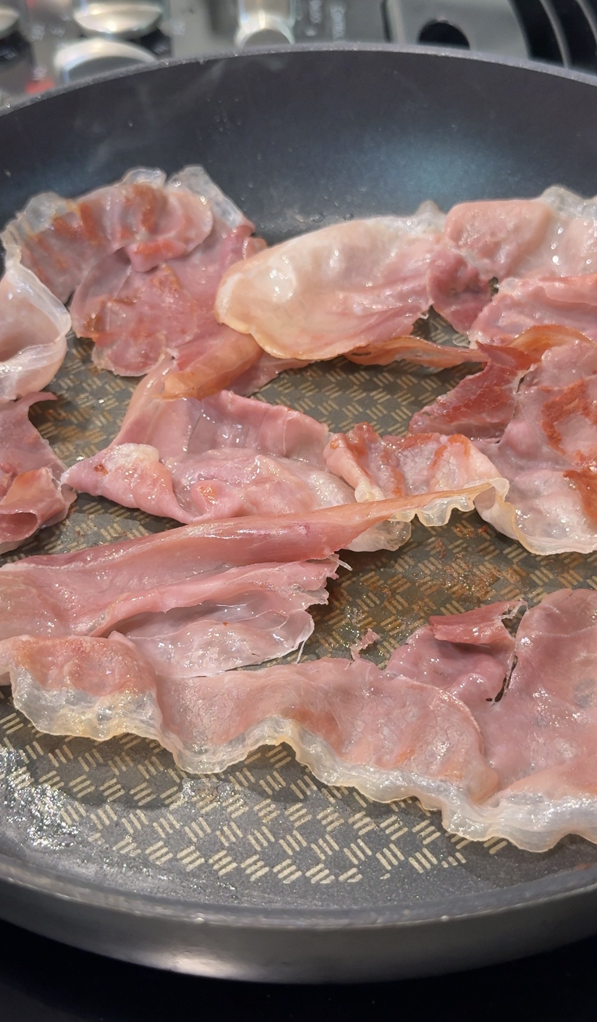 Several thin slices of prosciutto are spread out and cooking in a skillet with a textured, patterned surface.