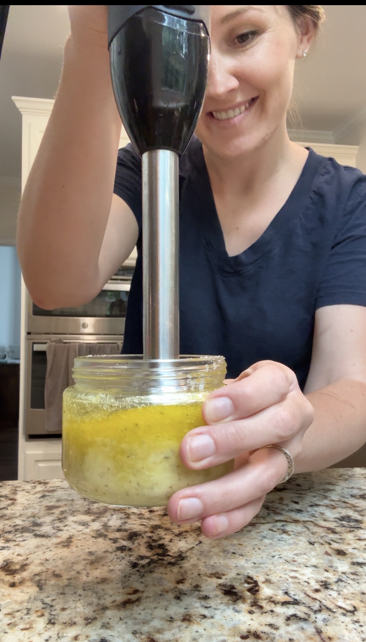 A woman in a navy shirt blends ingredients in a glass jar with an immersion blender on a granite countertop in a kitchen, smiling as she prepares food.