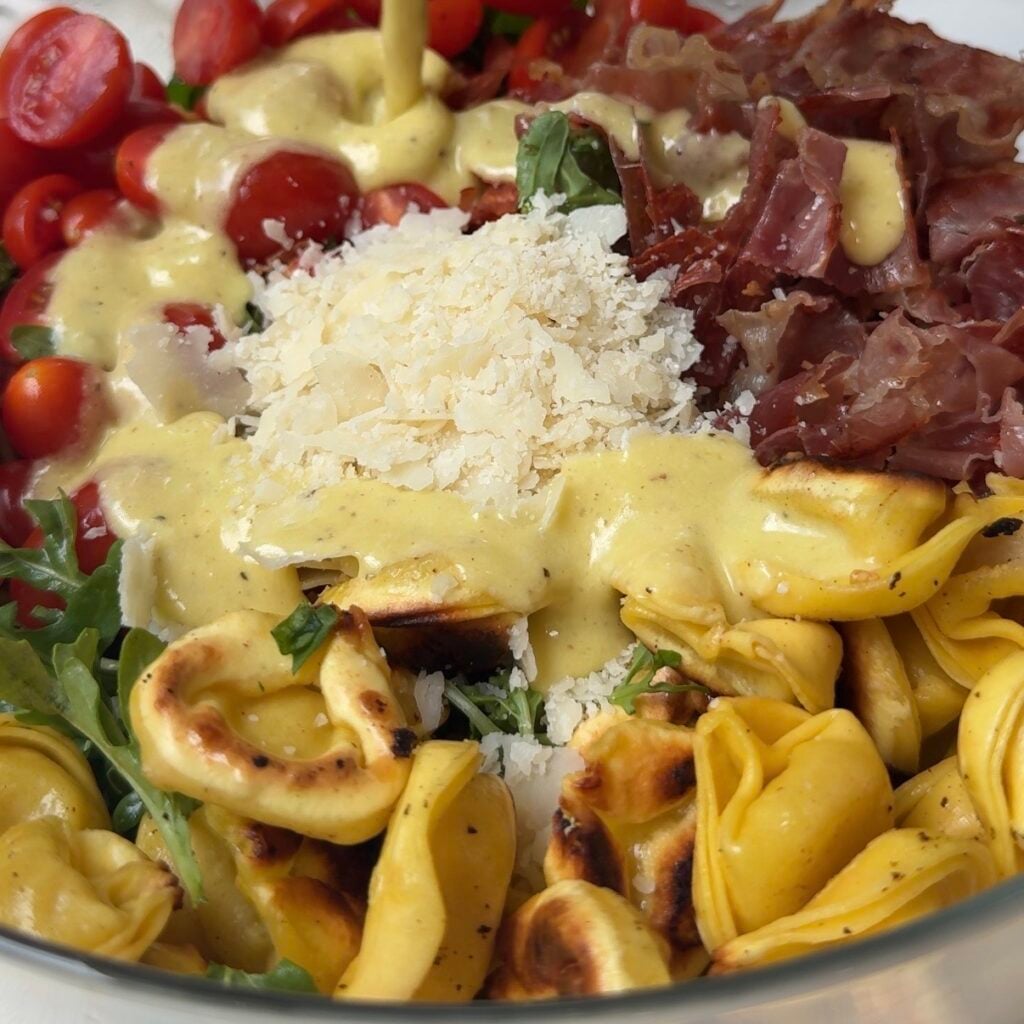 a bowl of salad with crispy tortellini, prosciutto, cherry tomatoes, parmesan and a dressing being over it