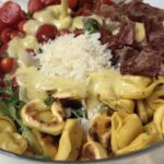 a bowl of salad with crispy tortellini, prosciutto, cherry tomatoes, parmesan and a dressing being over it