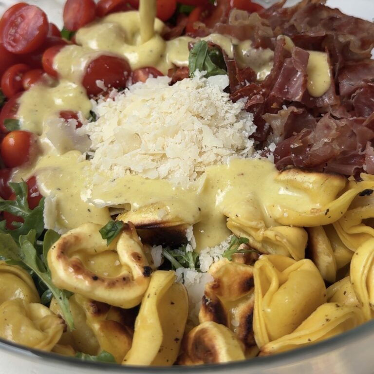 a bowl of salad with crispy tortellini, prosciutto, cherry tomatoes, parmesan and a dressing being over it