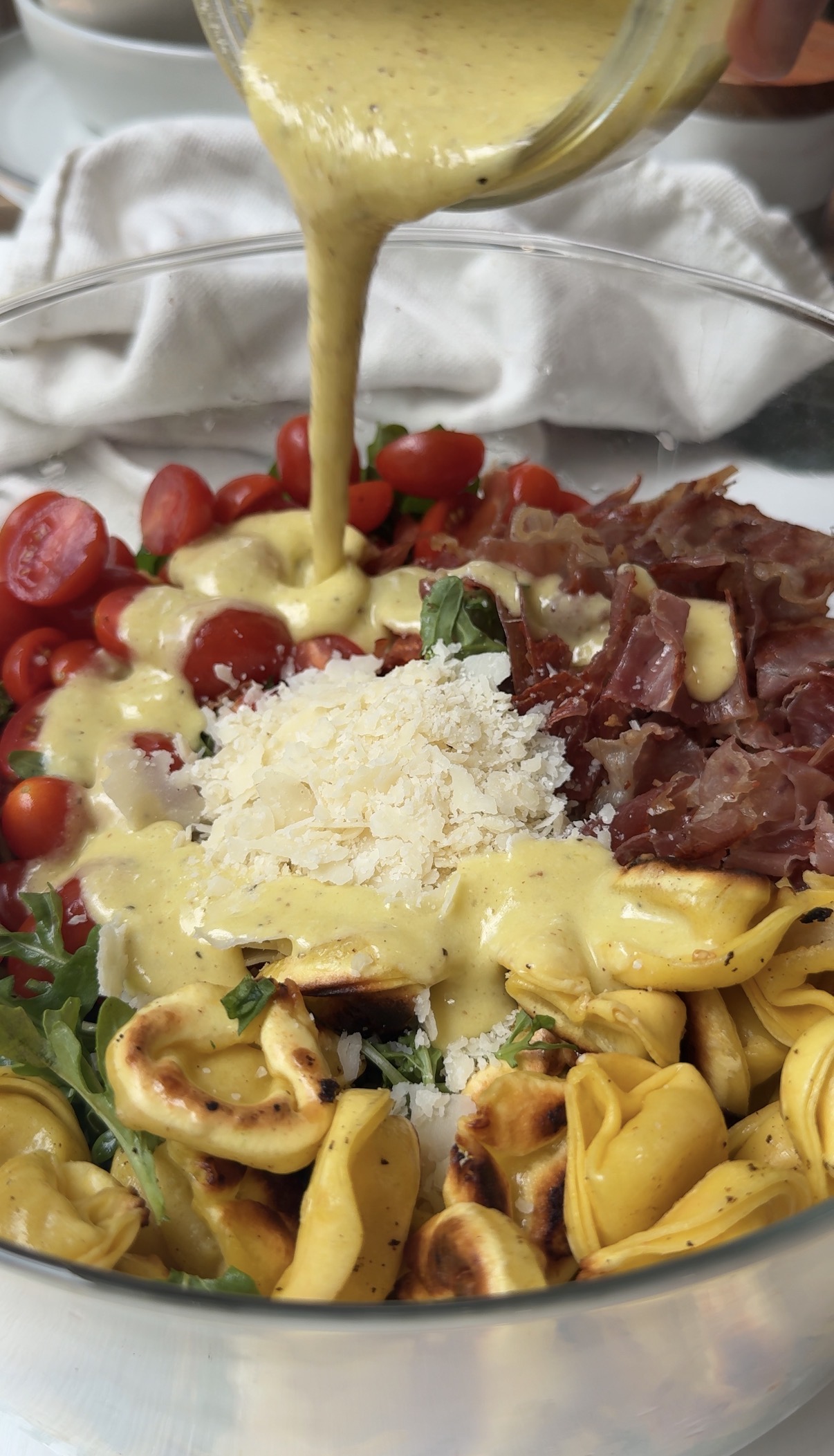 A close-up of a salad bowl with cherry tomatoes, prosciutto, tortellini, arugula, grated cheese, and creamy dressing being poured on top.