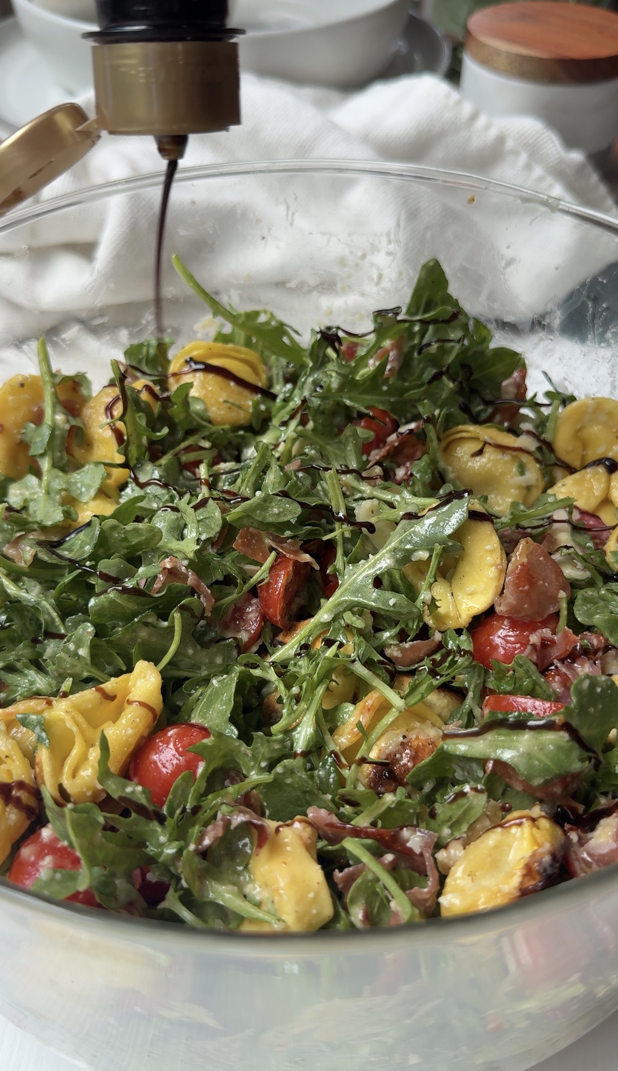 A glass bowl filled with a salad of mixed greens, tortellini pasta, cherry tomatoes, and shaved cheese, being drizzled with balsamic dressing.