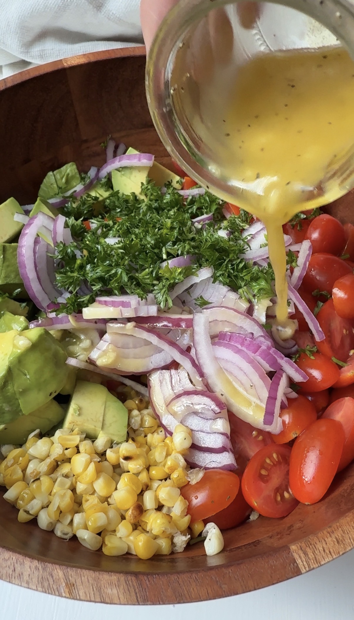 A wooden bowl filled with sliced red onions, cherry tomatoes, avocado chunks, roasted corn, and chopped parsley, with dressing being poured over the fresh salad.