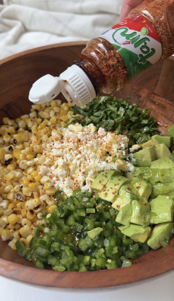A bowl with grilled corn, chopped cilantro, avocado, jalapeños, and crumbled cheese, with chili powder being sprinkled from a bottle on top.