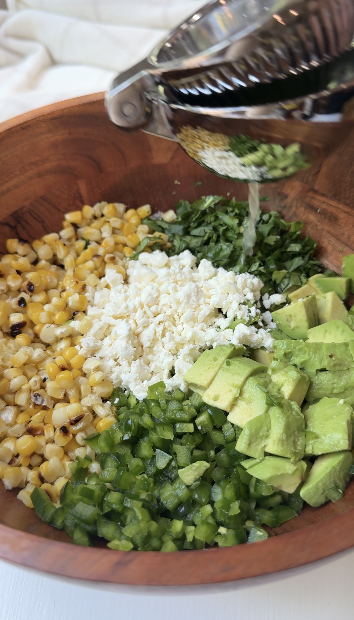 A wooden bowl filled with grilled corn, chopped cilantro, crumbled cheese, diced avocado, and chopped jalapeños, as lime juice is being squeezed over the ingredients.