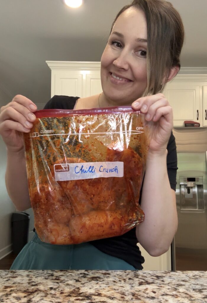 A woman in a kitchen holds a large ziplock bag filled with marinated chicken labeled Chilli Crunch. She is smiling at the camera. White kitchen cabinets and a stainless steel refrigerator are in the background.