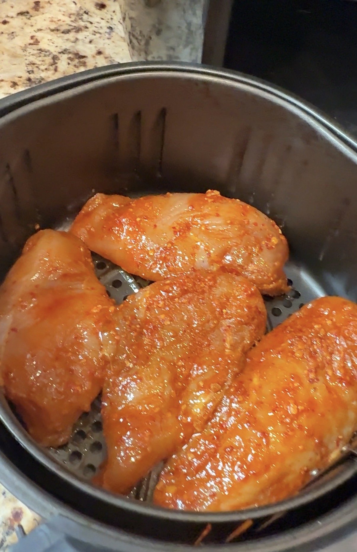 Four pieces of seasoned raw chicken breast are placed in an air fryer basket, ready to be cooked. The chicken is coated with a reddish spice blend.