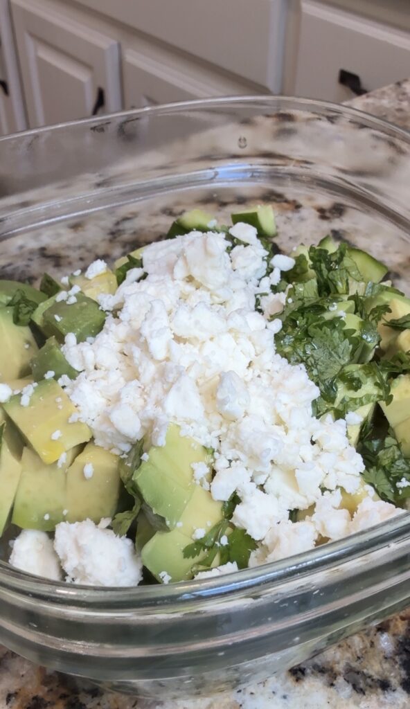 A glass bowl filled with diced avocado, chopped cilantro, and crumbled white cheese, sitting on a granite countertop.