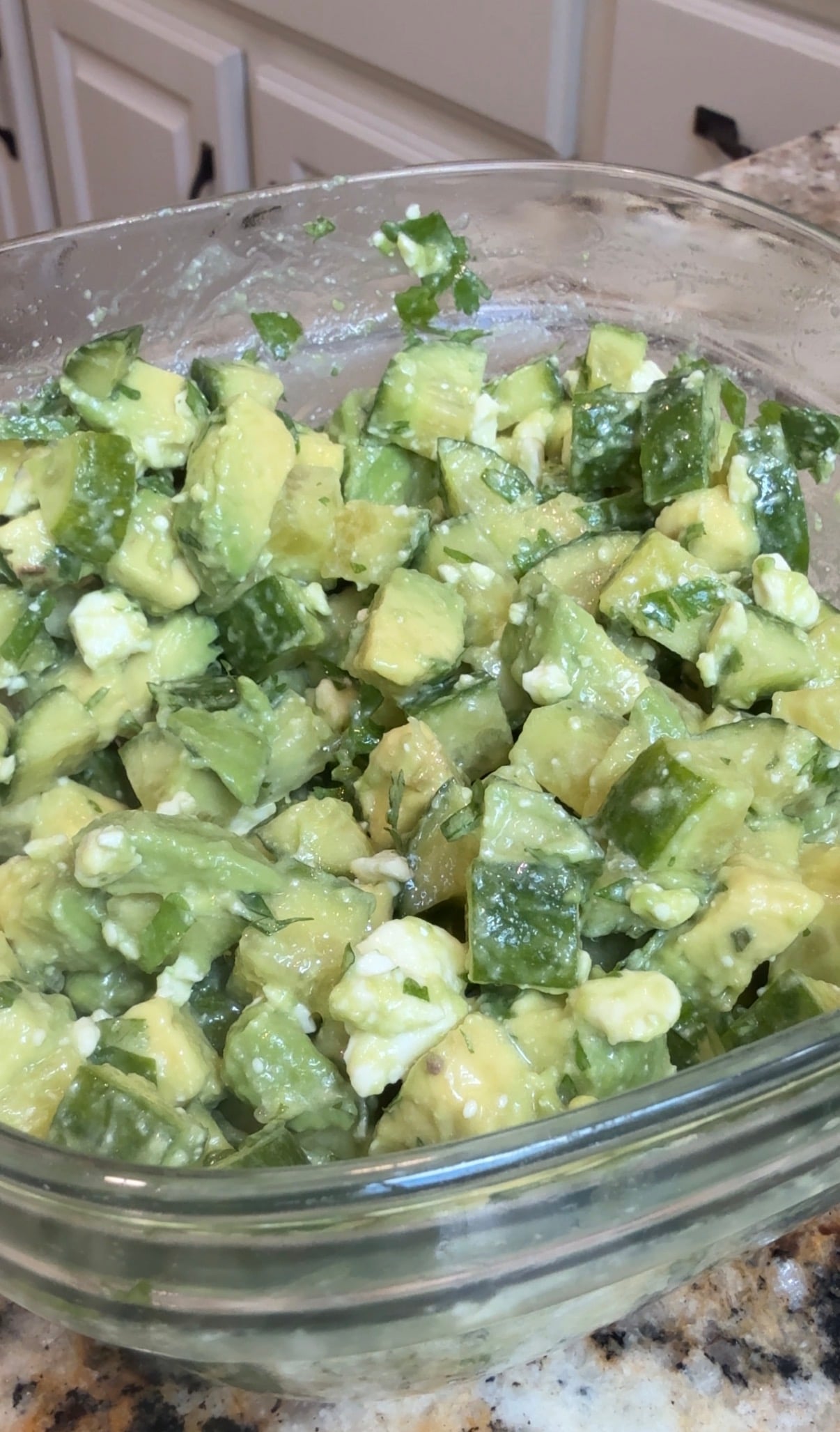 A glass bowl filled with a salad made of diced avocado, cucumber, crumbled feta cheese, and chopped herbs, sitting on a kitchen countertop.