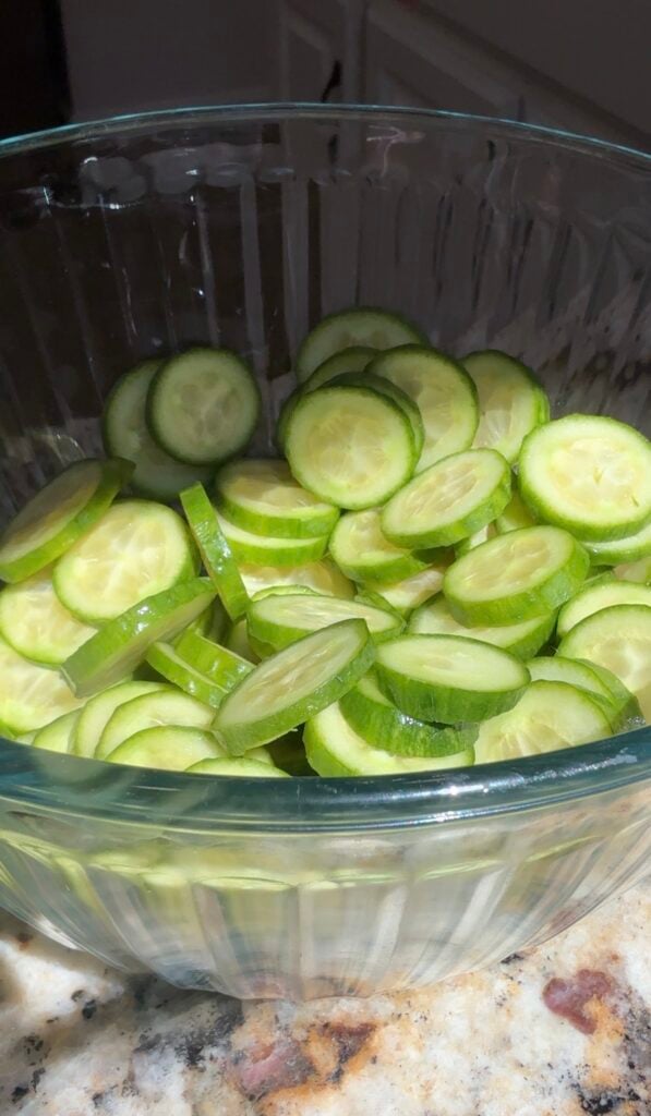 A clear glass bowl filled with thinly sliced cucumber rounds sits on a countertop, with sunlight highlighting the fresh, green color of the cucumbers.