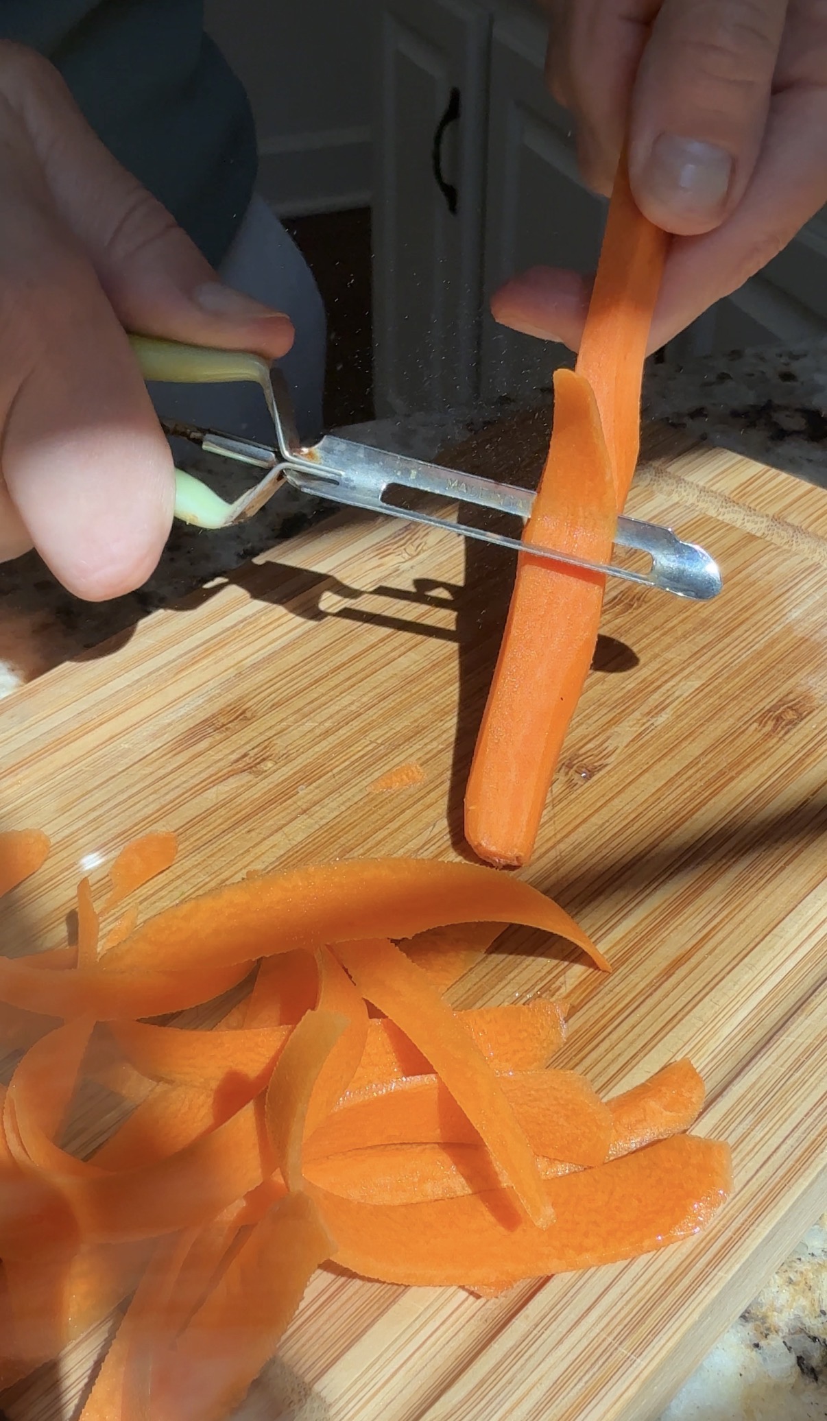 A close-up of a hand using a peeler to slice thin strips from a carrot over a wooden cutting board, with carrot peels scattered on the board.