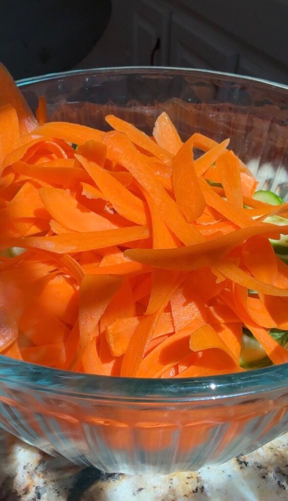 A glass bowl filled with thin, freshly peeled orange carrot ribbons, sitting on a kitchen counter in bright natural light.