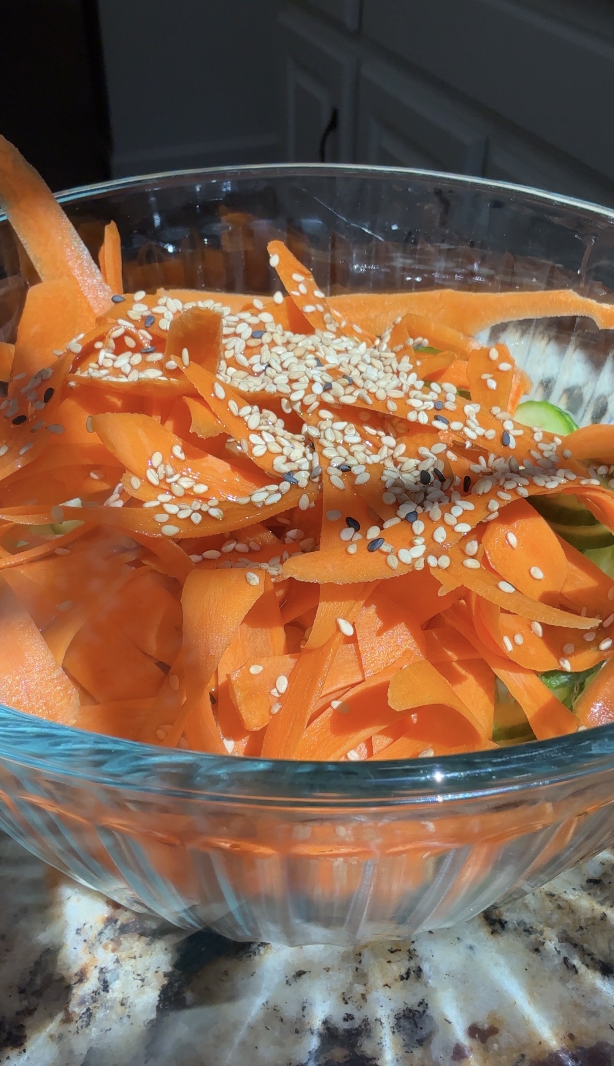 A glass bowl filled with thin carrot ribbons topped with white and black sesame seeds, with sunlight highlighting the vibrant orange color.
