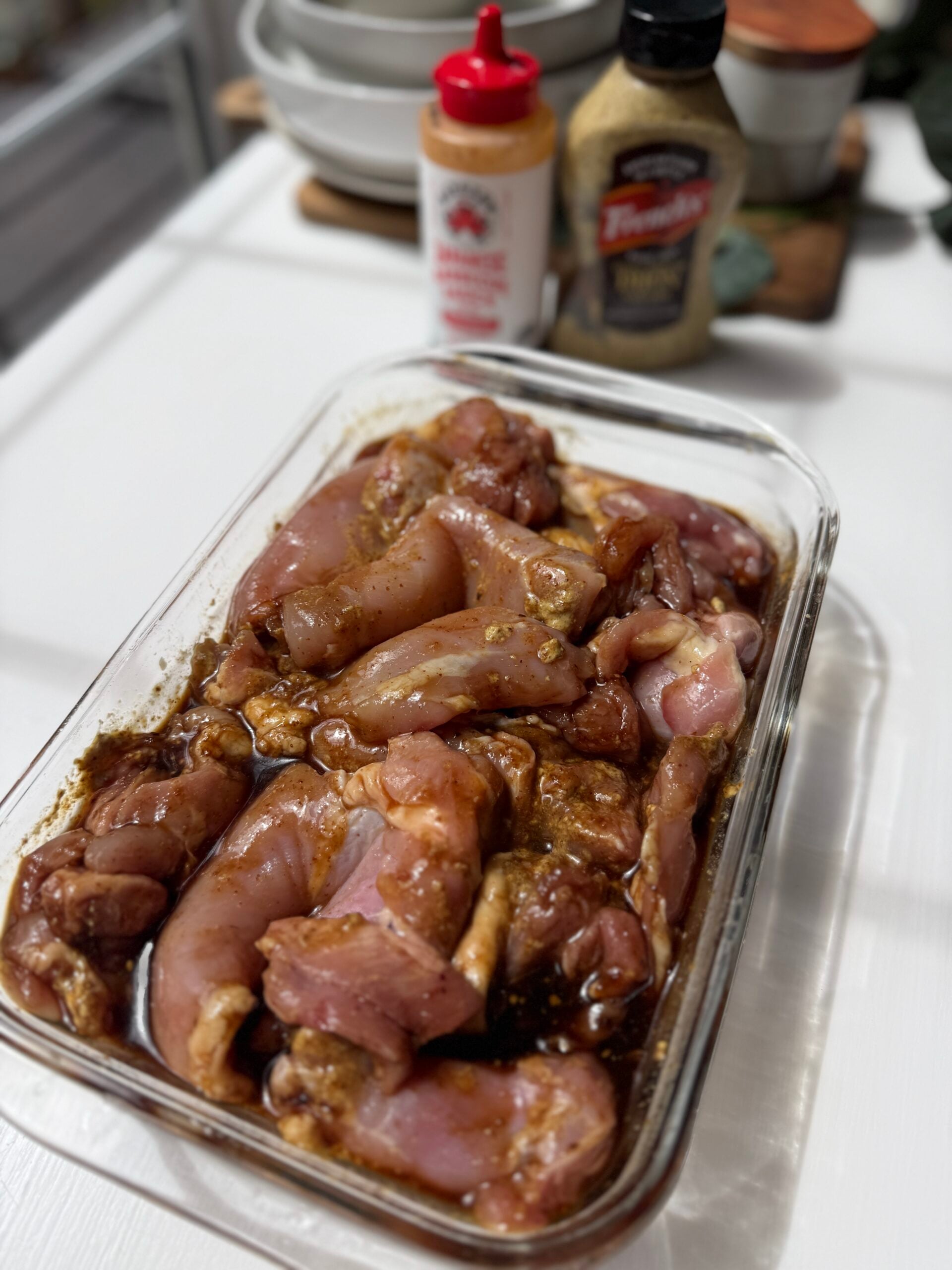 Raw chicken pieces marinating in a glass dish with spices and sauce. In the background, theres a bottle of Sriracha, a jar of Dijon mustard, and some stacked white bowls on a kitchen counter.