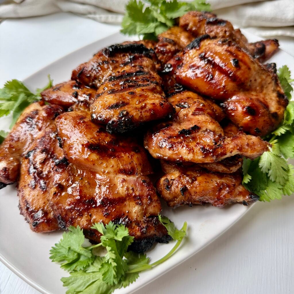 Grilled, glazed chicken thighs arranged on a white plate, garnished with fresh cilantro leaves, with a light-colored fabric in the background.