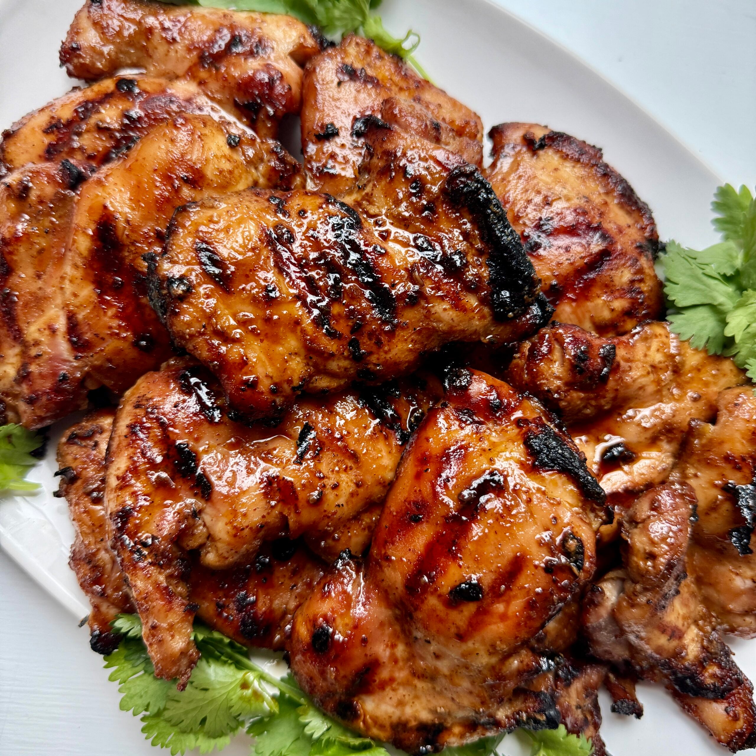 Grilled, glazed chicken thighs with a shiny, caramelized surface are arranged on a bed of fresh cilantro on a white plate.