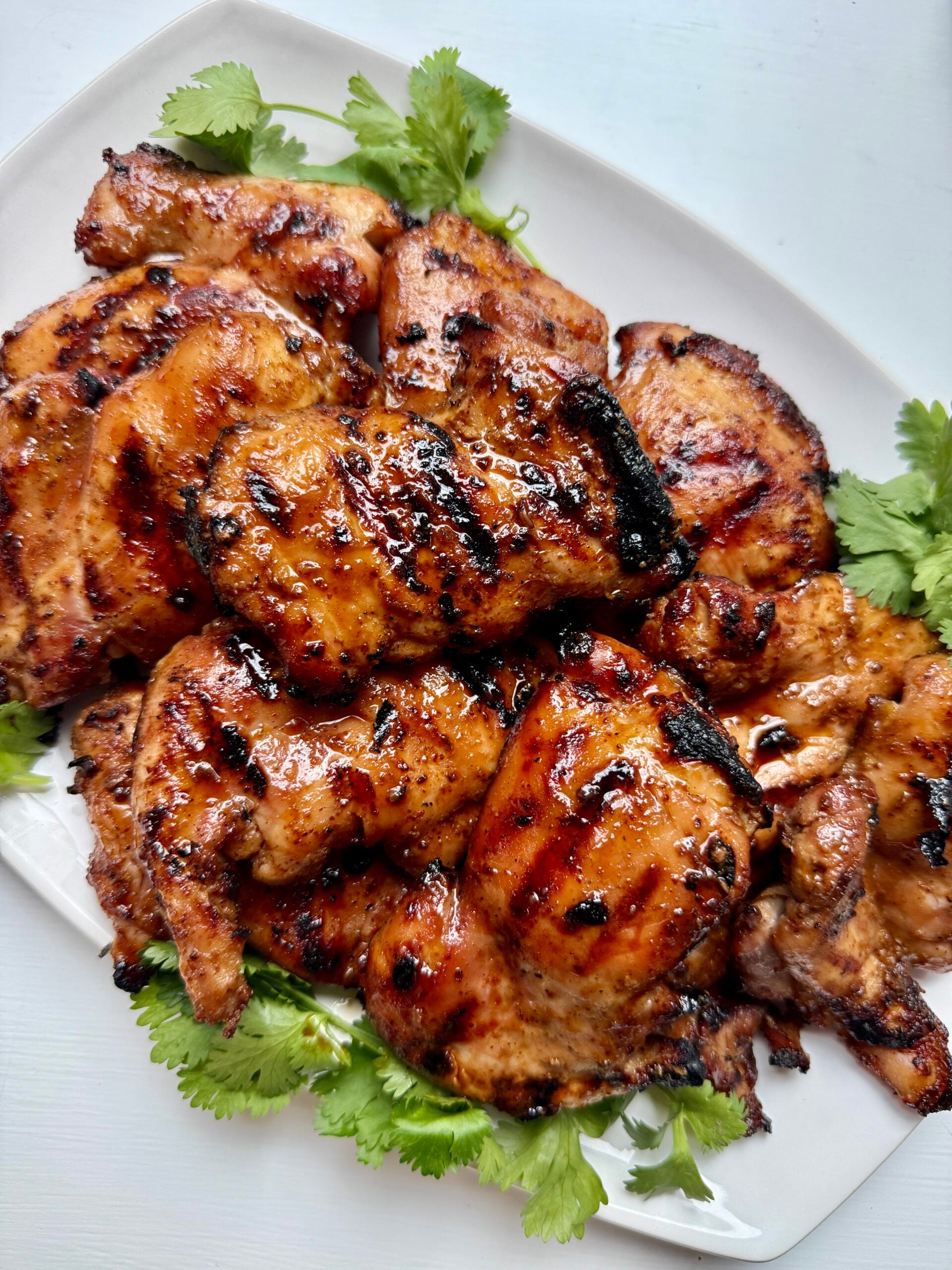 Grilled, glazed chicken thighs with a shiny, caramelized surface are arranged on a bed of fresh cilantro on a white plate.
