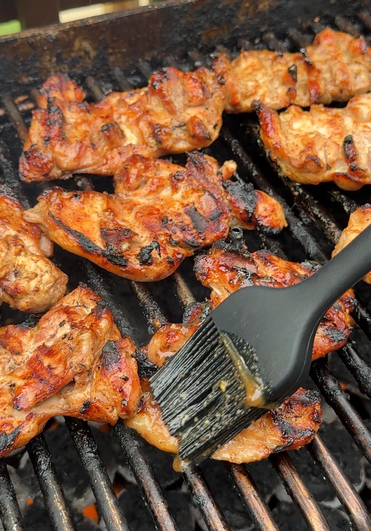 Grilled chicken pieces cooking on a barbecue grill, with a black silicone brush applying marinade or sauce to the meat.