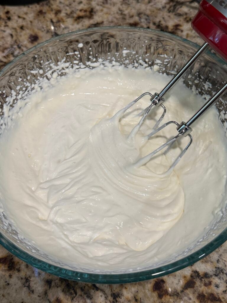 A glass bowl filled with whipped cream is being mixed with a handheld electric mixer, resting on a speckled kitchen countertop.