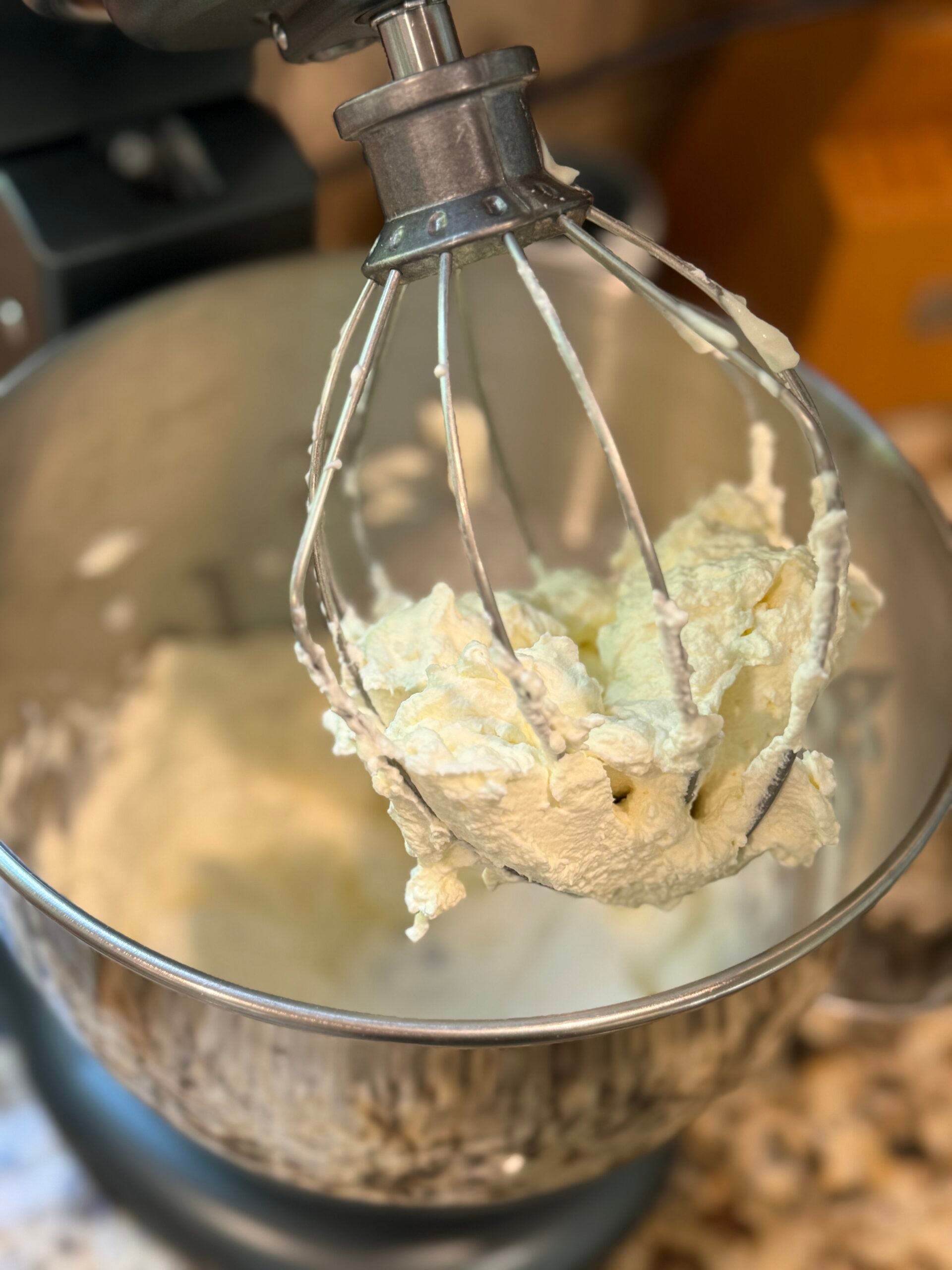 A close-up of a metal stand mixer with a whisk attachment covered in whipped cream or frosting. The mixing bowl below also contains some of the creamy mixture. The background shows a kitchen counter.