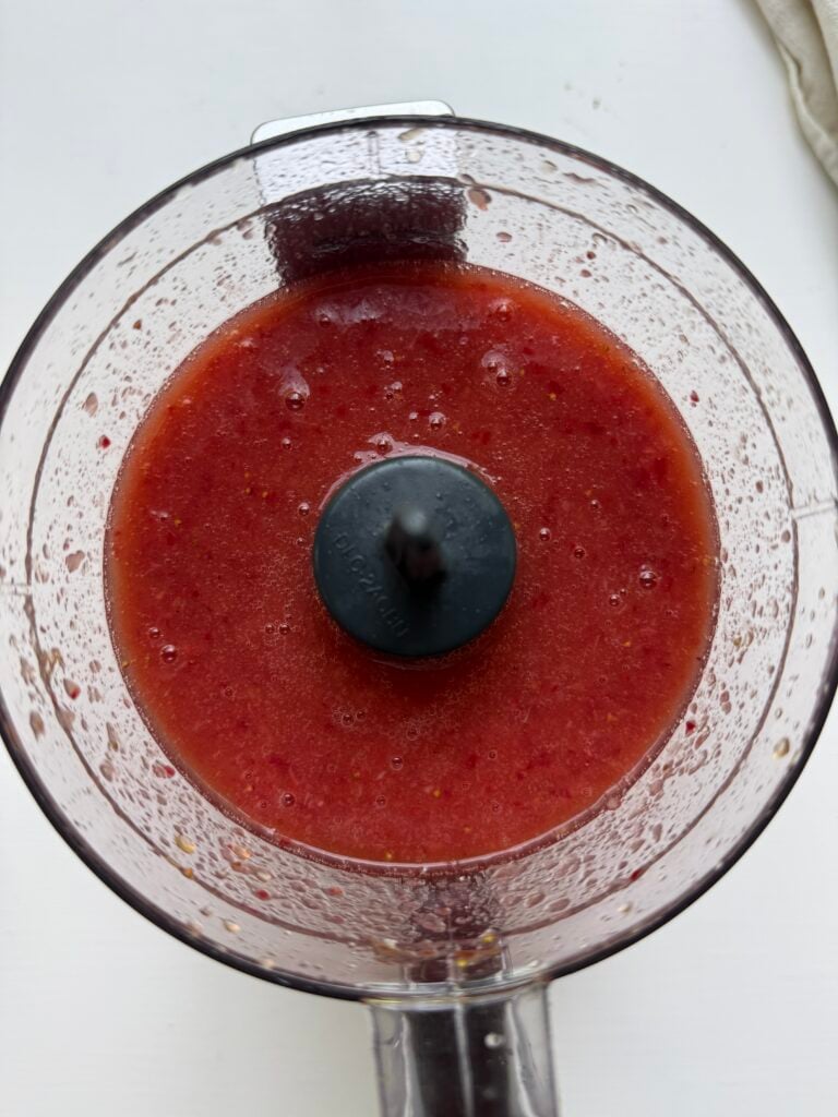 A food processor filled with a bright red, smooth blended mixture, likely a fruit puree, with droplets on the sides of the container.