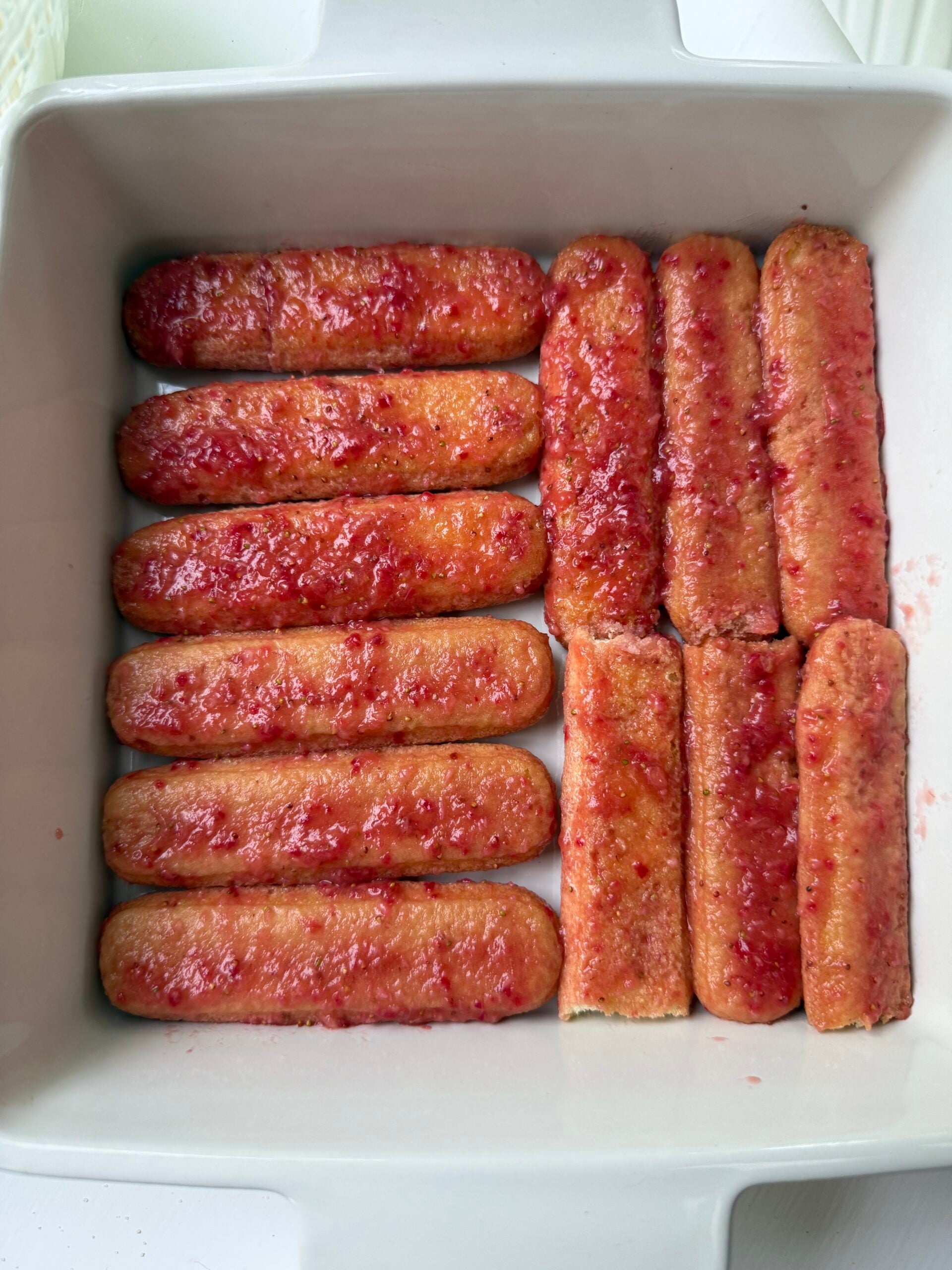 A white baking dish containing twelve ladyfinger biscuits, neatly arranged in two rows, each coated with a layer of red fruit syrup or compote.