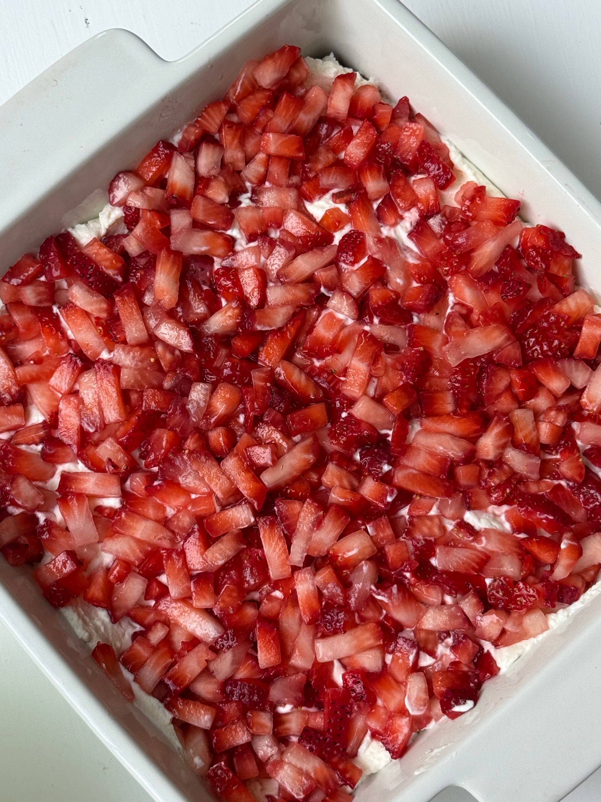 A square white baking dish filled with a layer of whipped cream or creamy mixture, topped with a generous layer of diced fresh strawberries.