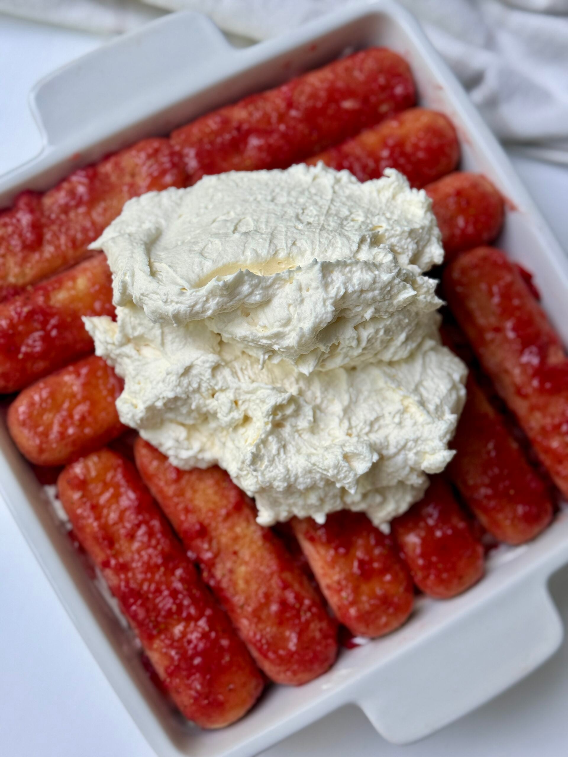 A white baking dish filled with ladyfinger biscuits soaked in a red liquid, topped with a generous mound of creamy white mascarpone mixture.