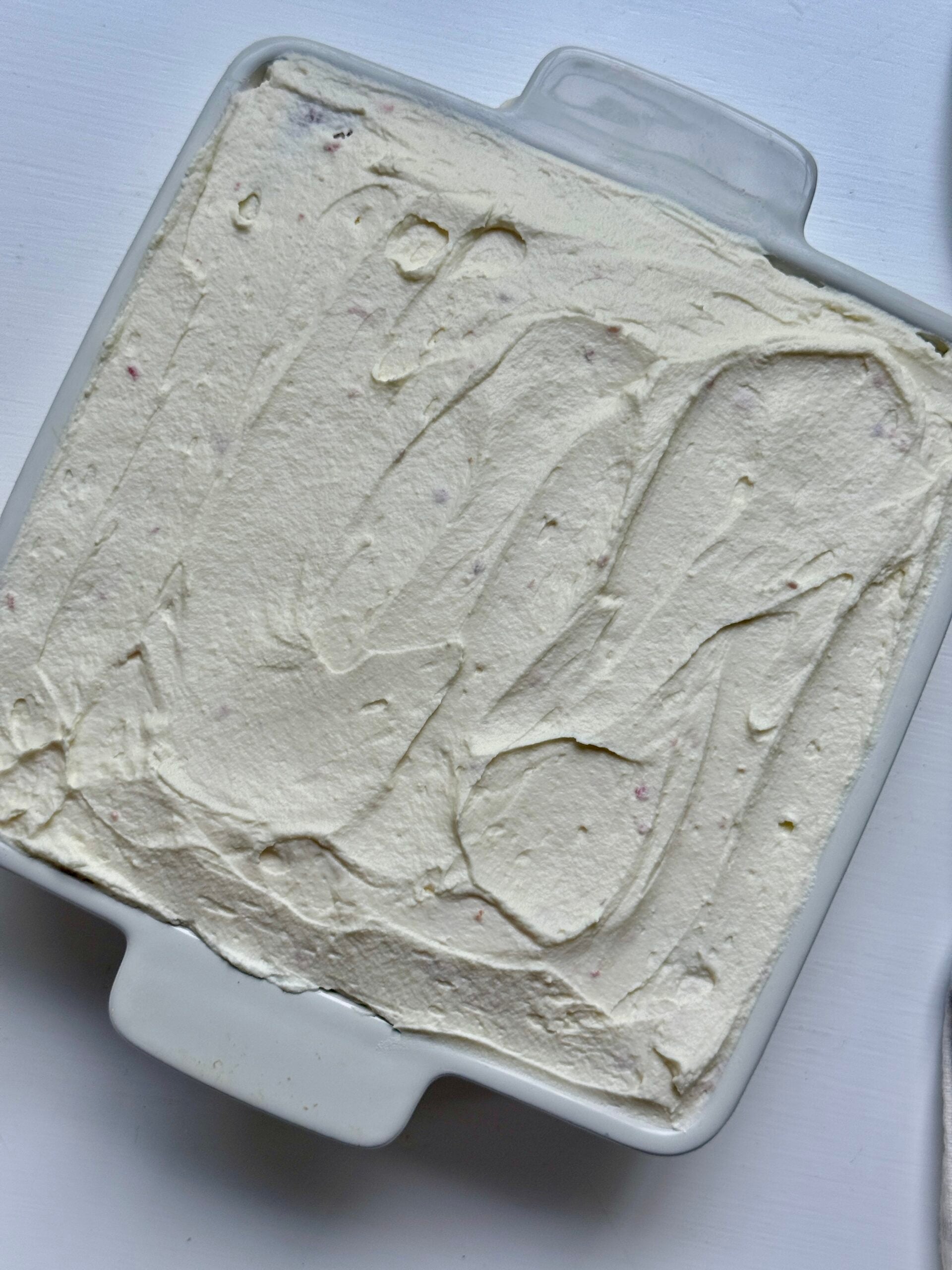 A square white baking dish filled with a creamy, smooth, light-colored mixture spread evenly across the surface, with visible swirl patterns on top. The dish is placed on a white background.