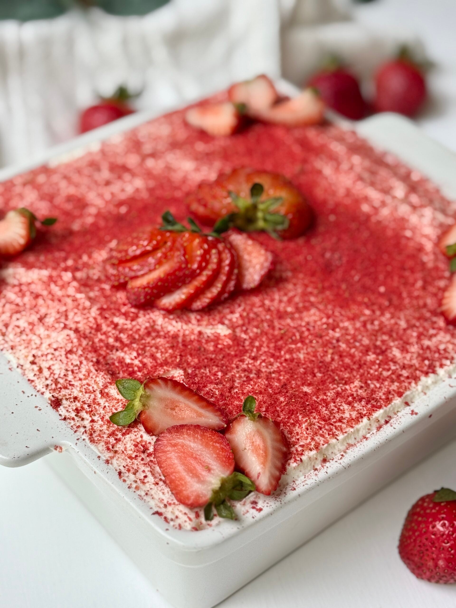 A white dish filled with a dessert topped with whipped cream, red strawberry powder, and fresh sliced strawberries, with whole strawberries placed around the dish on a white surface.