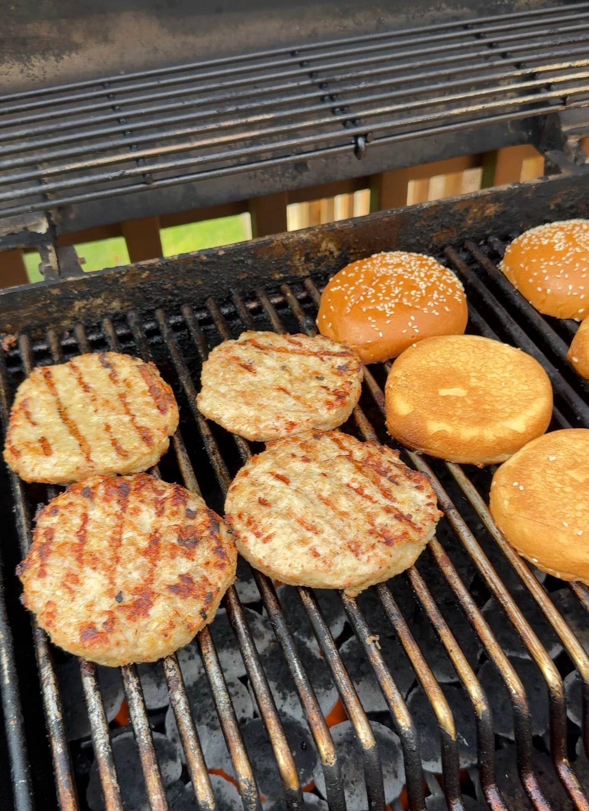 Four grilled burger patties and four toasted hamburger buns, two with sesame seeds, are cooking on a barbecue grill outside. The grill has visible char marks on the food and grates.