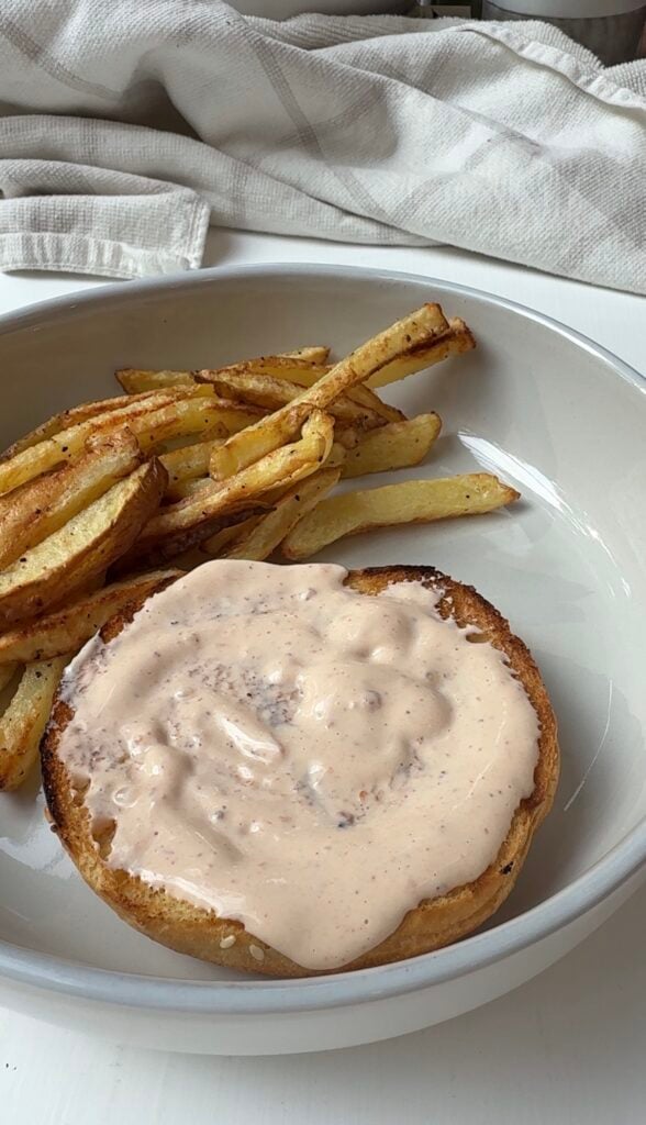A plate with golden, crispy French fries and an open-faced sandwich bun topped with creamy pink sauce, set on a white plate with a beige cloth in the background.