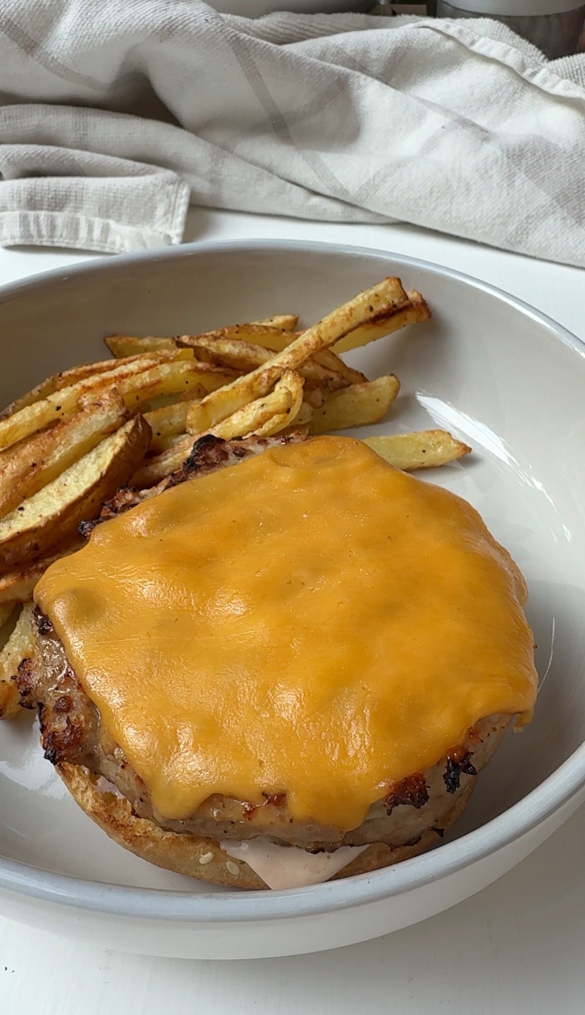 A plate with a cheeseburger topped with melted cheddar cheese and a side of golden-brown French fries, with a beige cloth napkin in the background.