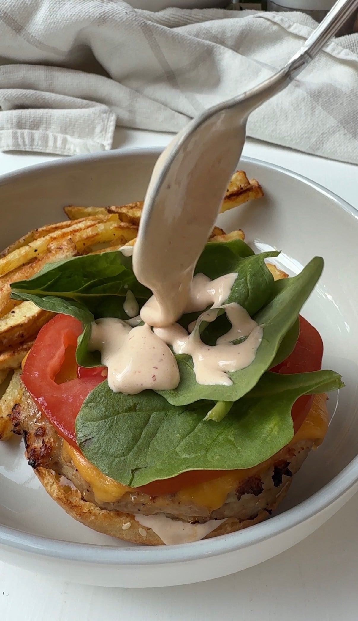 A spoon drizzles creamy sauce over fresh spinach and tomato on a burger patty with melted cheese, served in a bowl with golden French fries on the side.