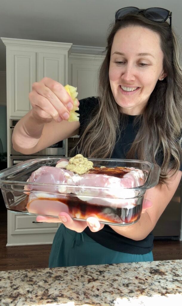 A smiling woman adds grated ginger to a glass baking dish filled with raw chicken and seasonings in a modern kitchen.