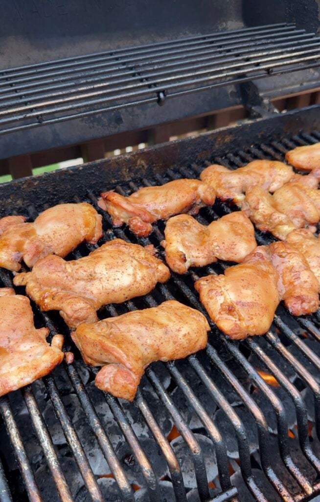 Several pieces of seasoned chicken are cooking on a grill, with grill marks visible on the meat. The grill grate is darkened from use, and the background shows the edge of the grill and part of a wooden deck.