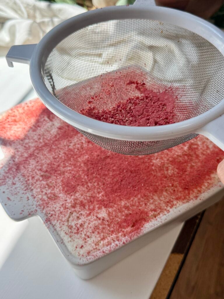 A hand holds a mesh strainer filled with powdered freeze-dried raspberries, sprinkling the pink powder onto a dish of creamy dessert below. Sunlight illuminates the scene on a white surface.