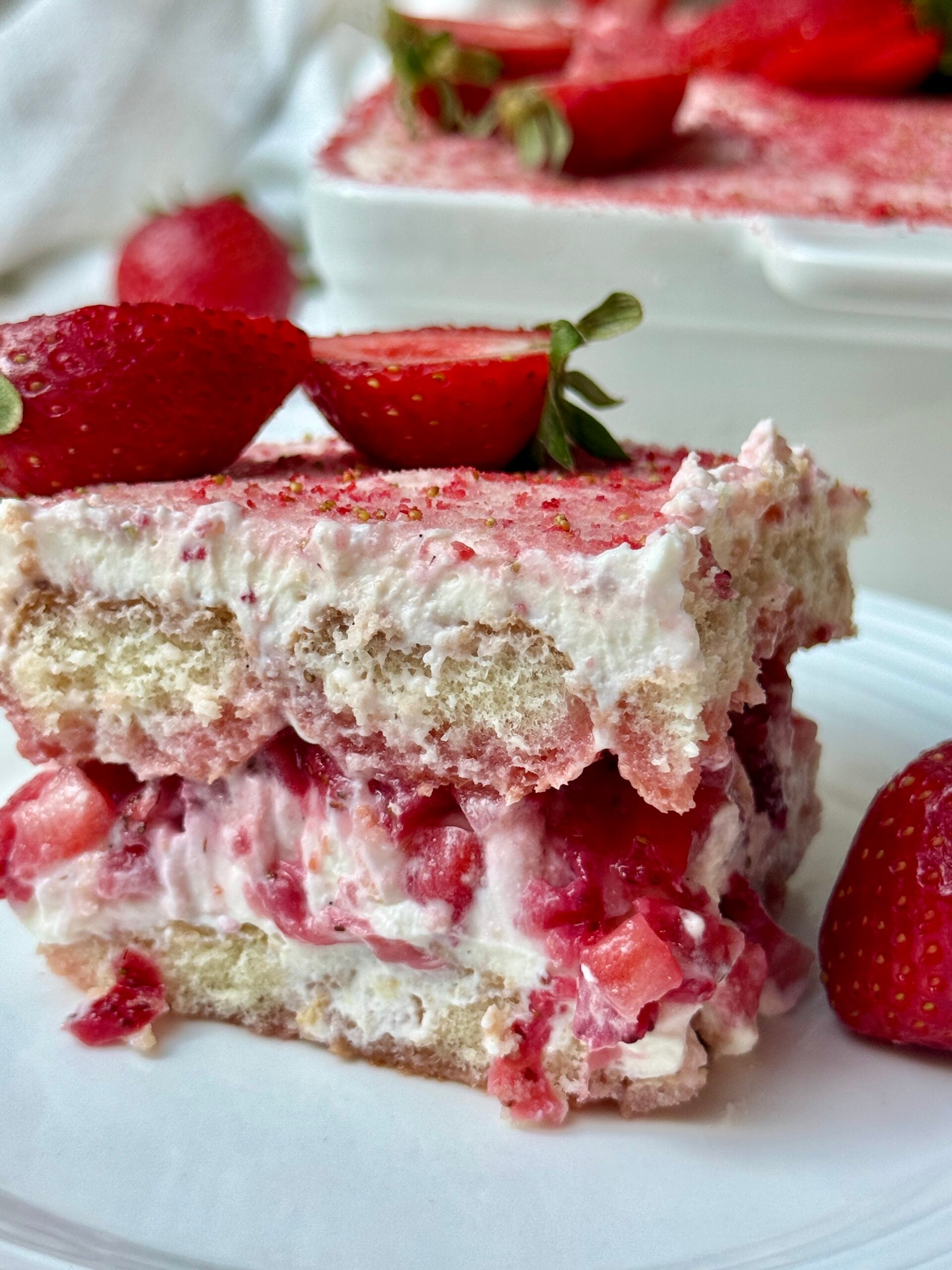A close-up of a slice of strawberry tiramisu on a white plate, topped with two halved strawberries, showing layers of cream, ladyfinger biscuits, and strawberry pieces, with whole strawberries nearby.