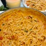 A large pan of creamy tomato pasta with spaghetti, garnished with fresh basil. In the background, a lit candle and a plate with a serving of the pasta are visible on a marble countertop.