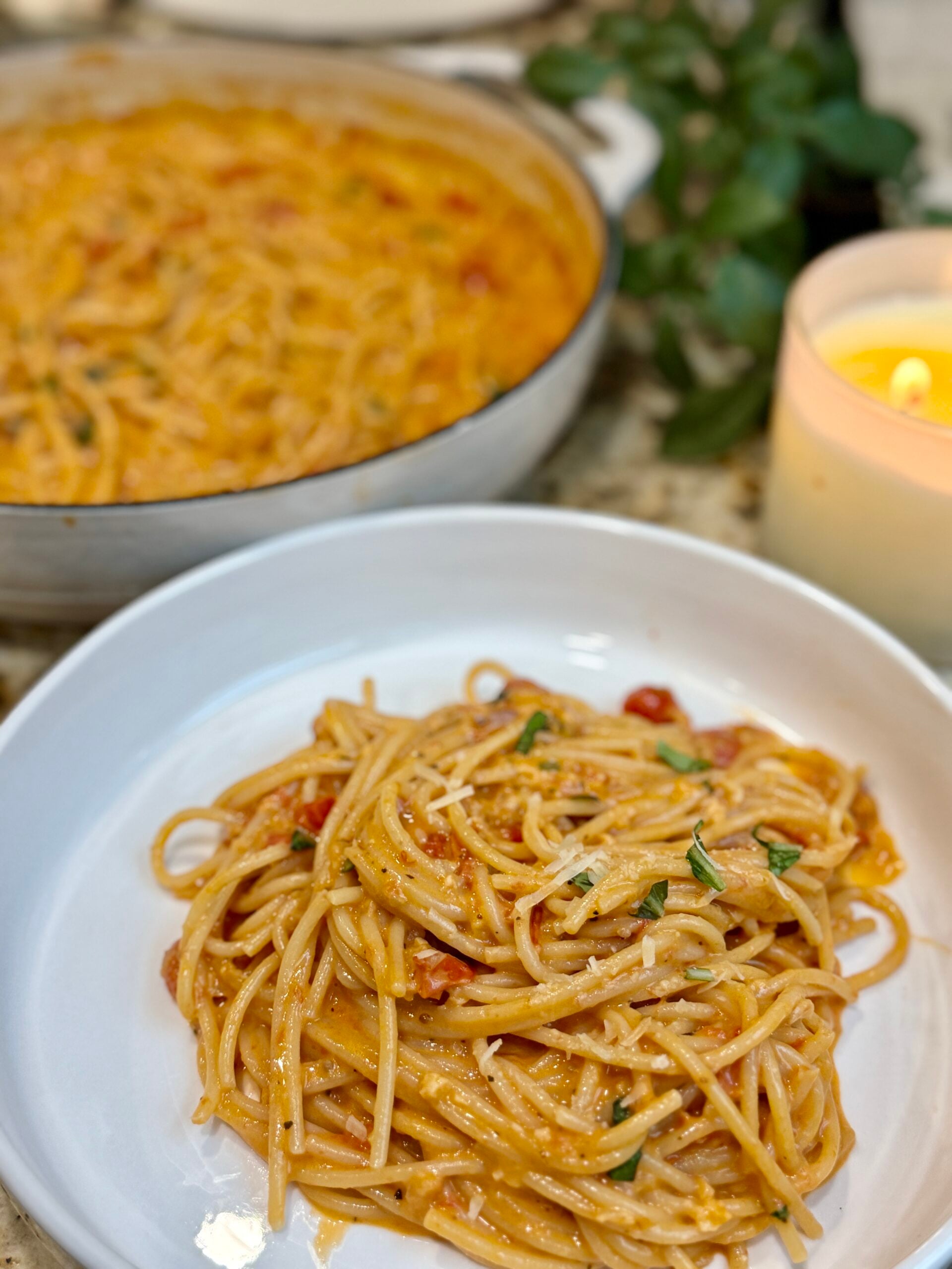 A plate of spaghetti with creamy tomato sauce and fresh herbs sits in the foreground, with a pan of more pasta and a lit candle visible in the background.