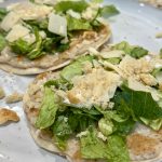 Two flour tortillas are being heated on a light-colored pan. The tortillas show slight browning in spots and appear to be lightly toasted. Some oil or melted butter is visible on the pans surface.
