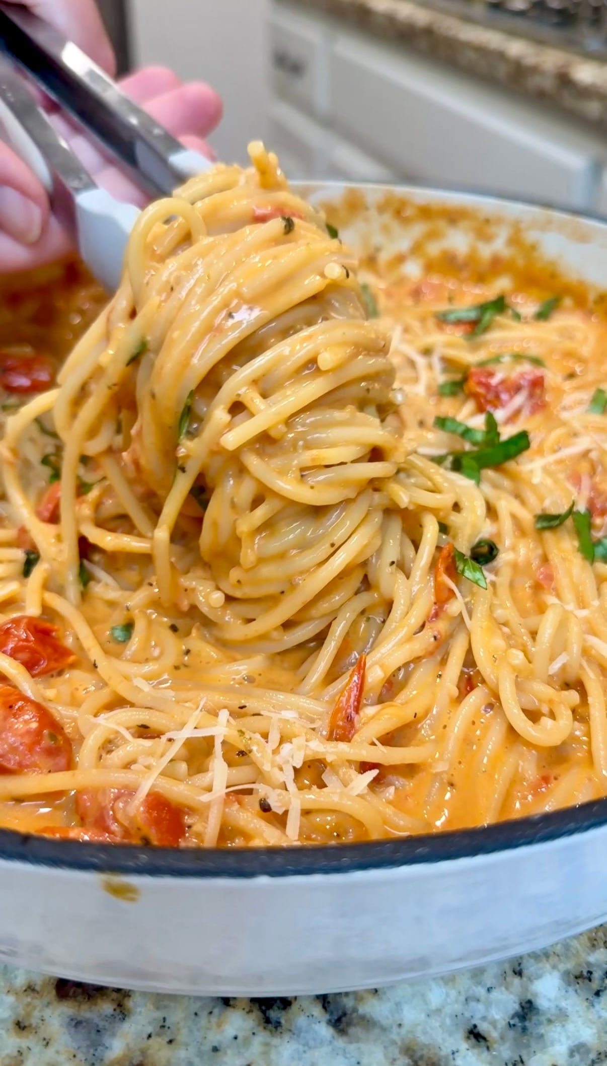 A close-up of creamy spaghetti being lifted with tongs from a pot, showing a rich sauce, cherry tomatoes, grated cheese, and chopped herbs.