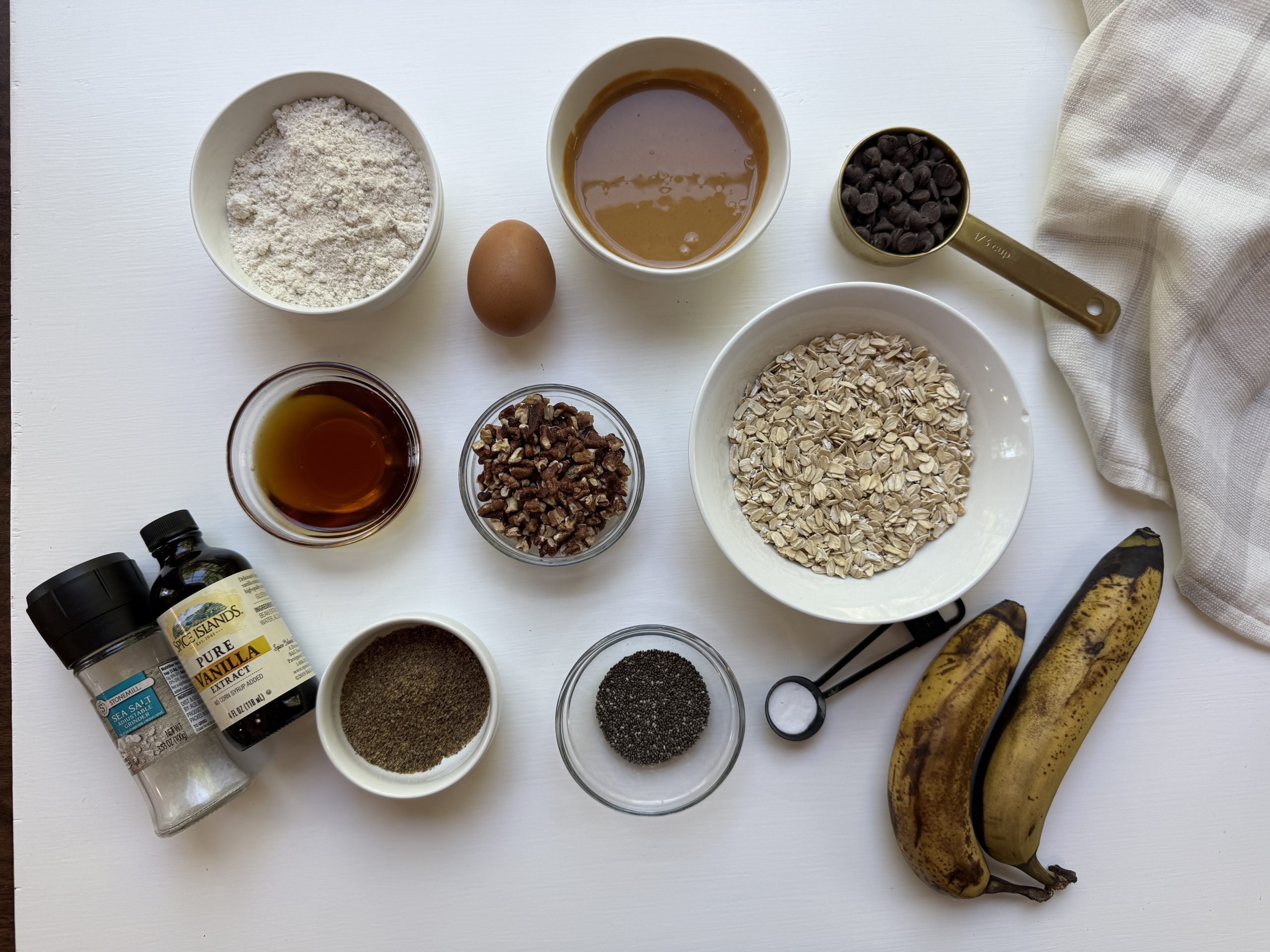Overhead view of baking ingredients on a white surface, including flour, an egg, peanut butter, chocolate chips, oats, pecans, maple syrup, bananas, chia seeds, flaxseed, vanilla, salt, and a kitchen towel.