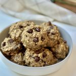 A white bowl filled with several oatmeal chocolate chip cookies, placed on a white surface with a blurred cloth and kitchen items in the background.