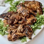 Grilled, marinated chicken thighs served on a white plate, garnished with fresh arugula leaves, on a light-colored surface.