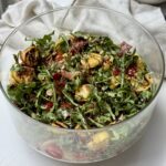 A glass bowl filled with a colorful salad of arugula, tortellini pasta, cherry tomatoes, prosciutto, and parmesan, drizzled with balsamic glaze, sits on a white surface with a white cloth in the background.