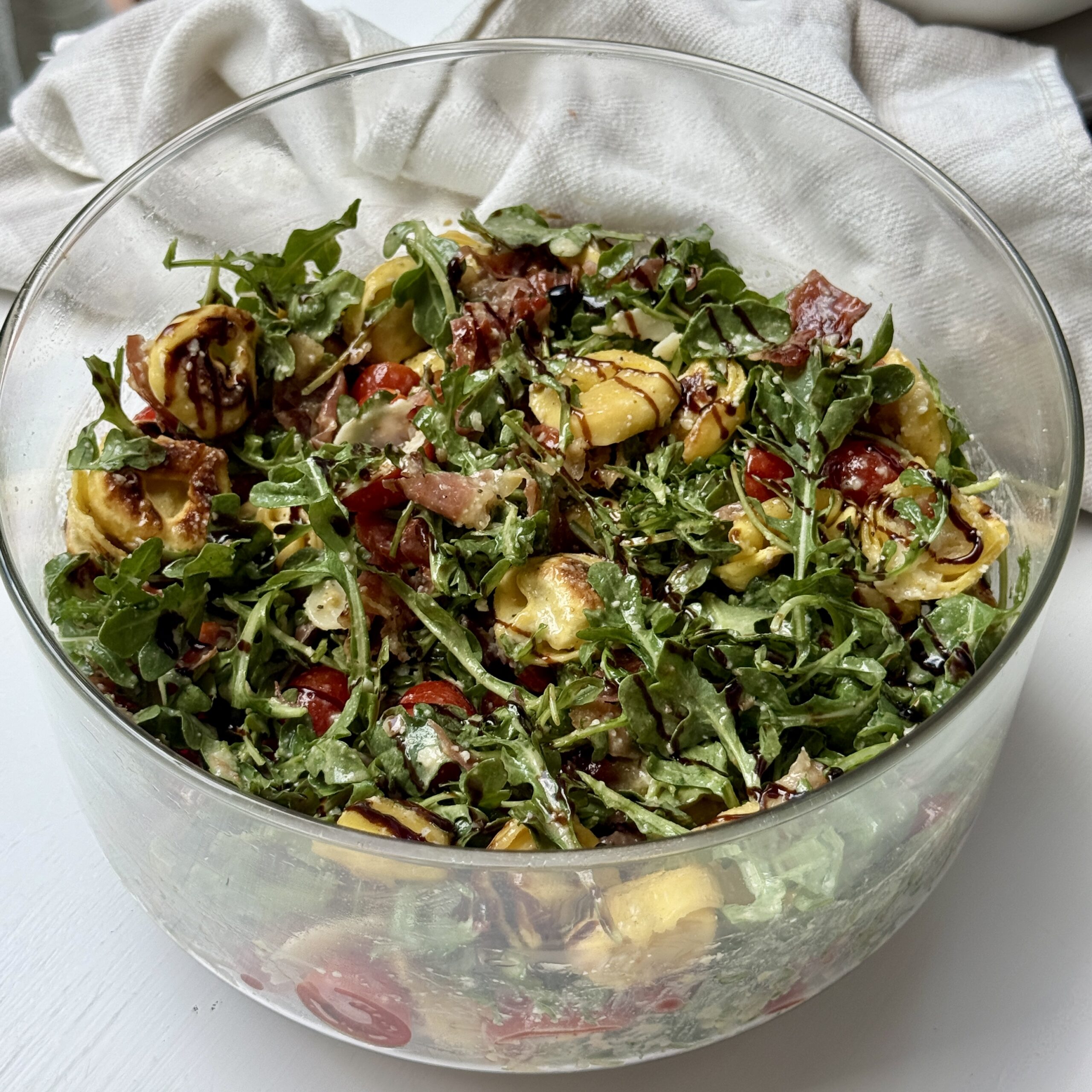 A glass bowl filled with a colorful salad of arugula, tortellini pasta, cherry tomatoes, prosciutto, and parmesan, drizzled with balsamic glaze, sits on a white surface with a white cloth in the background.