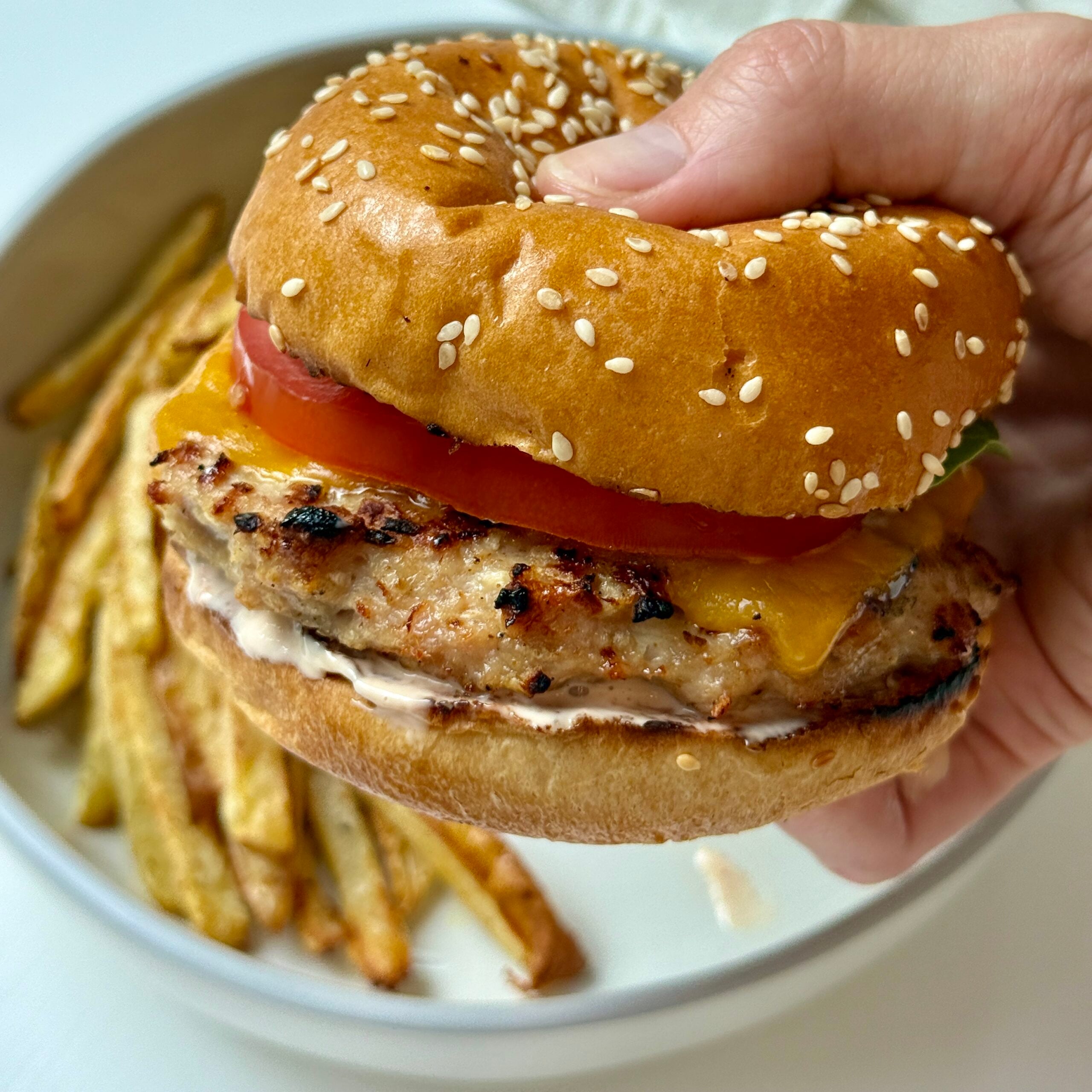 A hand holds a sesame seed bun sandwich with a grilled chicken patty, cheddar cheese, tomato, and sauce. In the background, a plate with French fries is visible.