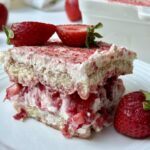 A slice of strawberry tiramisu layered with cream and strawberries sits on a white plate, garnished with a halved strawberry on top and a whole strawberry next to it. More strawberries and the dessert dish are in the background.